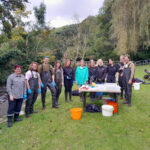A group of DCRT staff and volunteers standing together beside a river during a Riverfly training session, with equipment laid out on a table in front of them.
