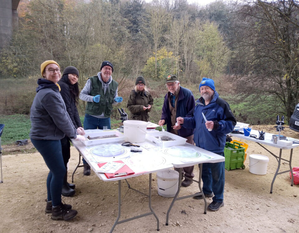 A group of volunteers standing around tables set up with trays and sampling equipment during an extended Riverfly survey under a viaduct.
