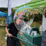A person creating a giant soap bubble at a DCRT outdoor event, standing in front of information stalls and display tents.