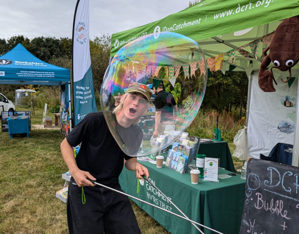 A person creating a giant soap bubble at a DCRT outdoor event, standing in front of information stalls and display tents.