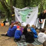 Children sitting on a blanket in a woodland clearing during an outdoor River Guardians lesson, with an instructor speaking in front of a large map backdrop and activity materials scattered nearby.