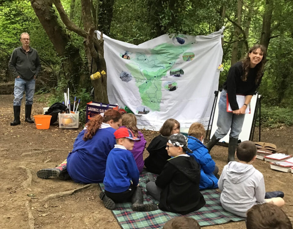 Children sitting on a blanket in a woodland clearing during an outdoor River Guardians lesson, with an instructor speaking in front of a large map backdrop and activity materials scattered nearby.