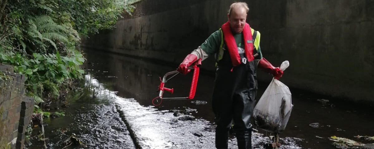 A volunteer wearing protective gear removing litter from a river channel.
