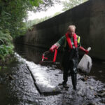 A volunteer wearing protective gear removing litter from a river channel.