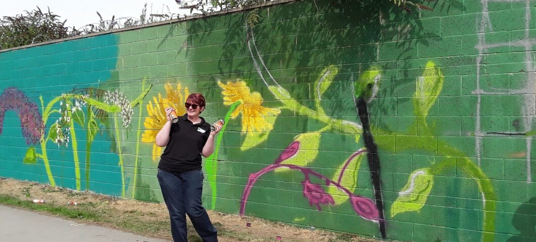 A volunteer standing beside a colourful mural created as part of a riverside community art project.
