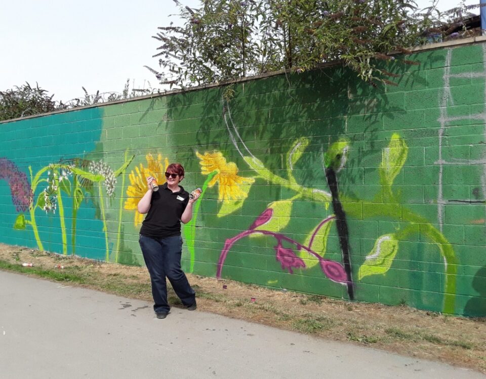 A volunteer standing beside a colourful mural created as part of a riverside community art project.