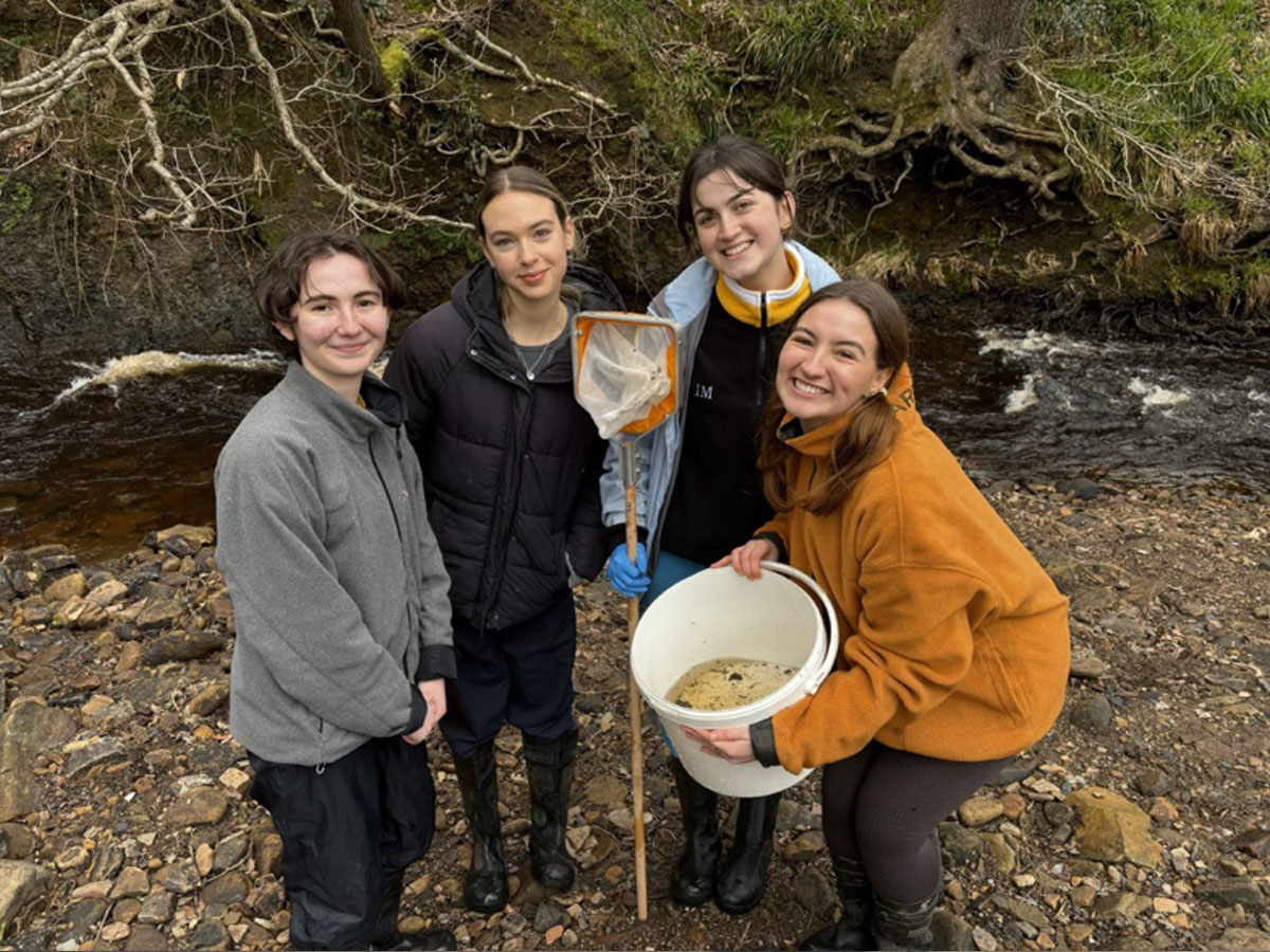 Group of young volunteers smiling beside a river while holding a sampling net and bucket during a survey activity.
